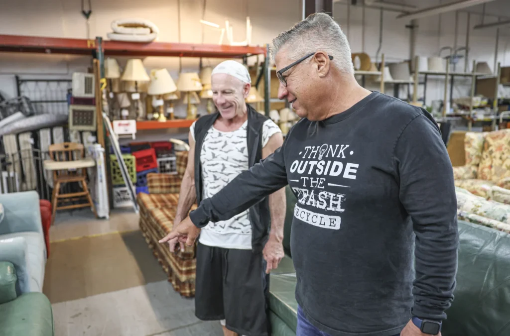 Two men stand in a warehouse filled with furniture and household items. One man points at a piece of furniture.
