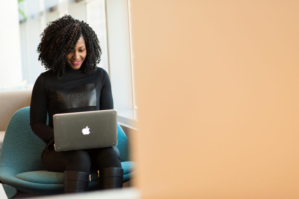A sitting African-American woman smiles while typing on the laptop resting on her knees. She is wearing a black sweater, black pants and black boots.