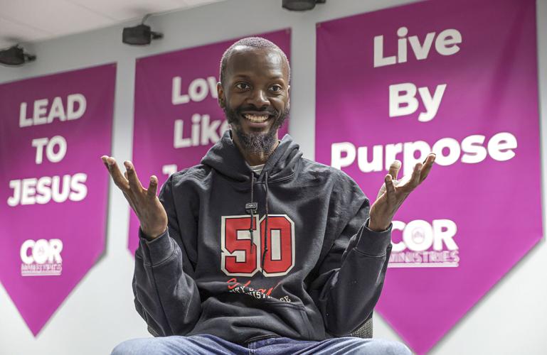 An African-American man sits in front of purple religious banners. He is wearing a black sweatshirt with the number 50 on the front and blue jeans. He has short black hair and a beard. His hands are raised in gesture and