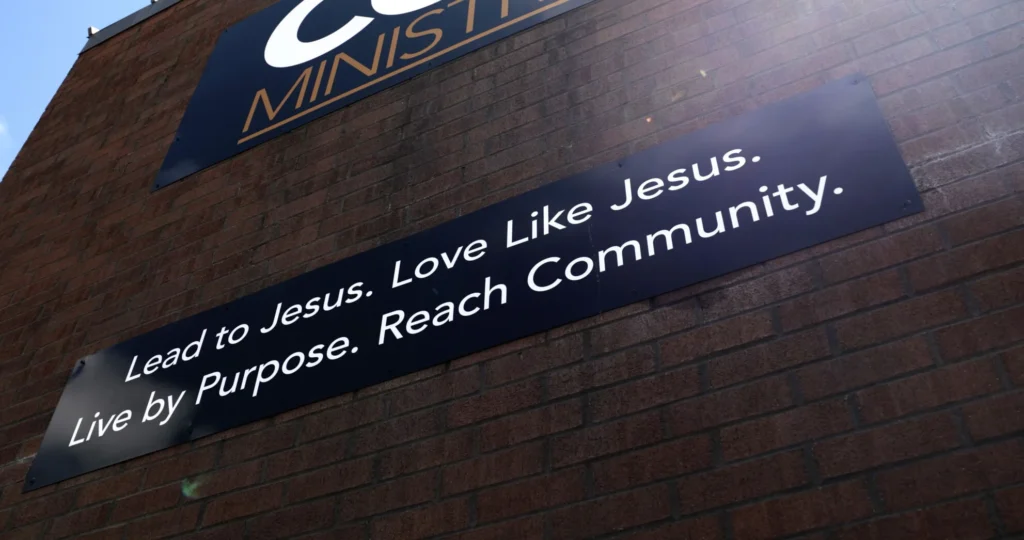 A brick wall is shown with a black sign reading "Lead to Jesus. Love like Jesus. Live by purpose. Reach Community" in white letters.
