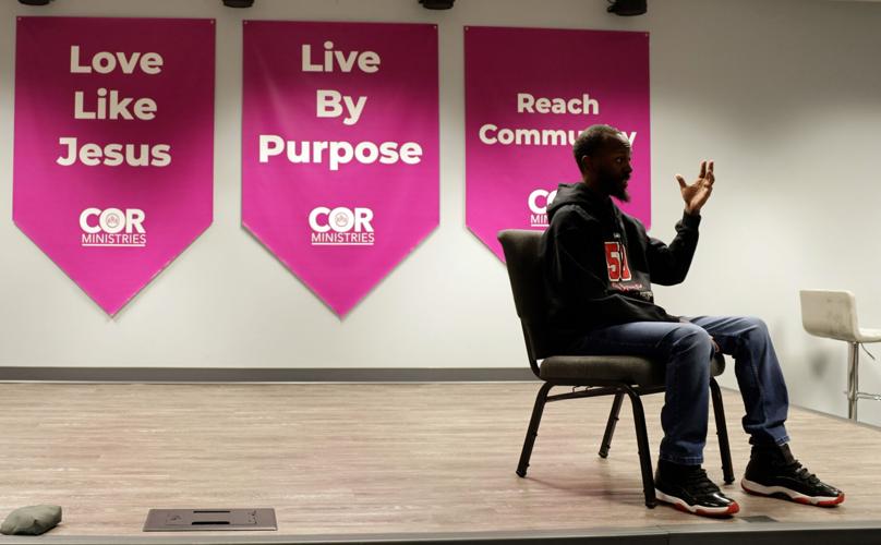 An African-American man with black hair and a beard sits in a chair on a raised platform with purple religious banners in the background. He is wearing a black hooded sweatshirt, blue jeans and black tennis shoes.