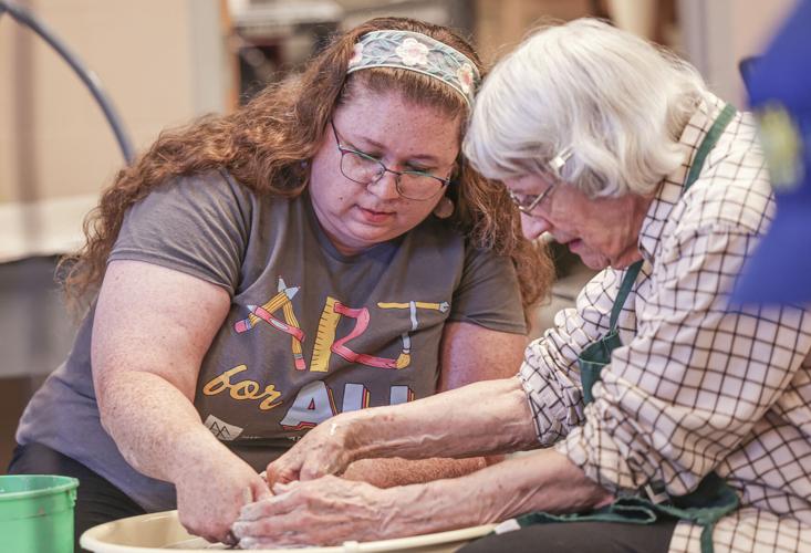 Two women sit at a pottery wheel with both of their hands working on a piece of pottery. The woman on the left has long red hair held back with a bandana and wears wire-rimmed glasses and a grey T-shirt which says "Art for All." The woman on the right has grey hair and wears wire-rimmed glasses and a white and brown patterned shirt rolled up to the elbows.