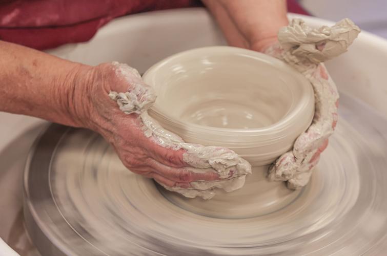A woman's hands work to create a clay pot on a pottery wheel. The hands are coated with clay slip and cup a bowl shape.