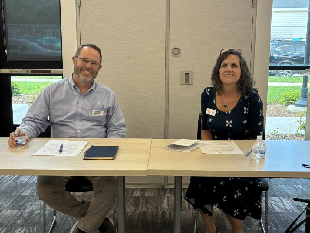 A white adult man and woman sit at a presentation table. The man, who has a beard, wears wire-rimmed glasses, a light blue oxford shirt and tan pants. The woman at right wears red glasses pushed up in her hair, a dark blue dress featuring a white pattern.