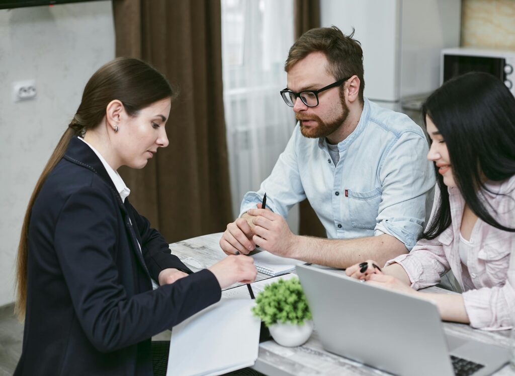 A woman with long brown hair pulled back into a ponytail sits facing a man and woman. The woman on the left wears a white business shirt and blue jacket and uses a pen to point to some paper on the table between her and the couple. The man has short brown hair, a beard and mustache, and wears black-framed glasses and a light blue shirt rolled to his elbows. The woman at this side has long black hair and wears a pink long sleeved shirt over a white shirt. In the background is a window with dark brown curtains and sheers.