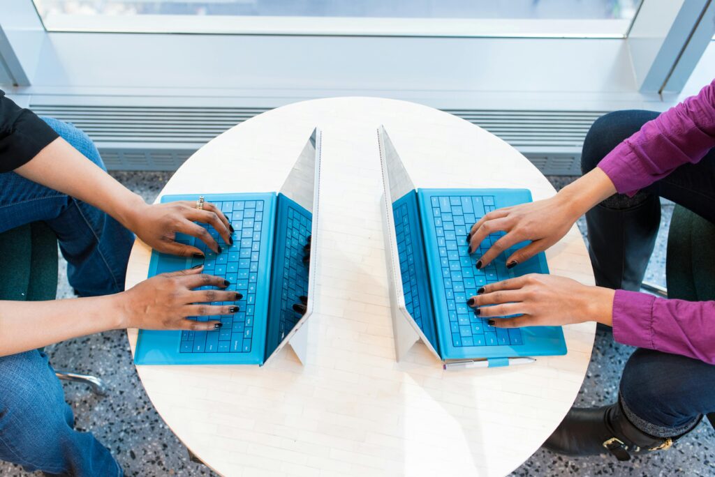 Two open blue laptops are shown on a small round table facing away from each other. At each keyboard, a woman sits with her hands placed on the keyboard. Both women are wearing black nail polish. The woman on the left wears blue jeans and a black top, while the woman on the right wears dark jeans, a purple shirt and black boots.