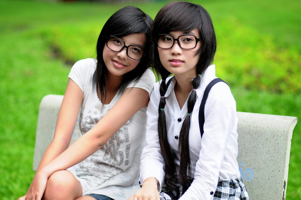Two young women of Asian heritage sit on a bench with grass in the background. The woman on the left has shoulder-length black hair and wears black-framed glasses, a white T-shirt with slogans on the front. The woman on the right has long black hair with braids down her front and wears black-framed glasses, a white dress shirt with a black and white plaid skirt with black suspenders. Both women look directly at the camera.