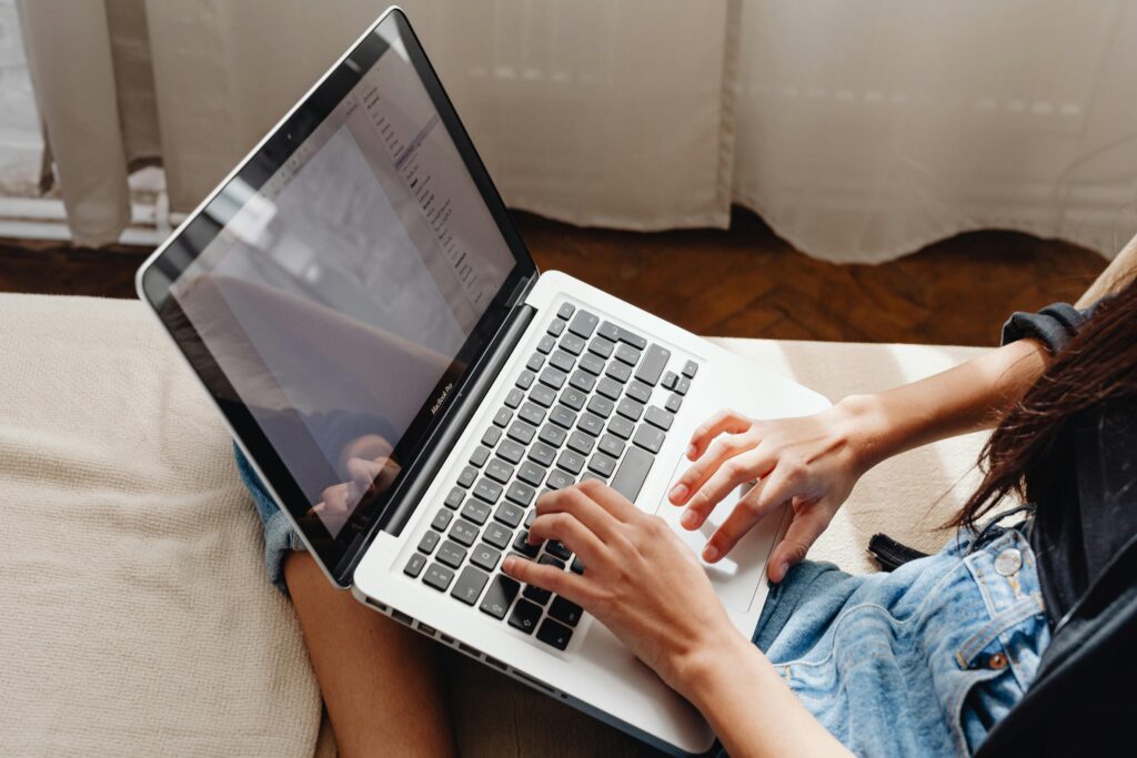 A laptop is shown sitting on a young woman's lap as she sits near a window with sheer curtains.
