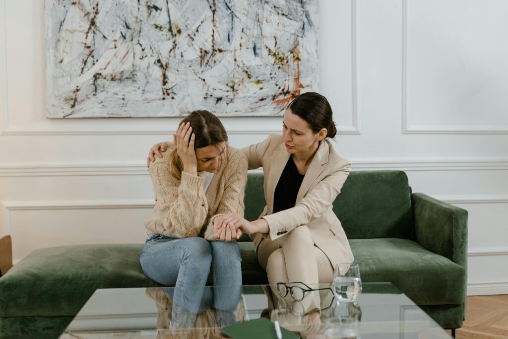 Two women sit on a green couch. The woman on the left holder her head in her hand while the woman on the right holds her other hand and has her arm around the woman on the left. The woman on the left is wearing a beige sweater and jeans, while the woman on the right is wearing a beige suit with a black shirt underneath the jacket. A glass coffee table sits in the foreground and it has a pair of eyeglasses and a glass of water resting on it. Behind the women, an abstract painting hangs on a white wall.