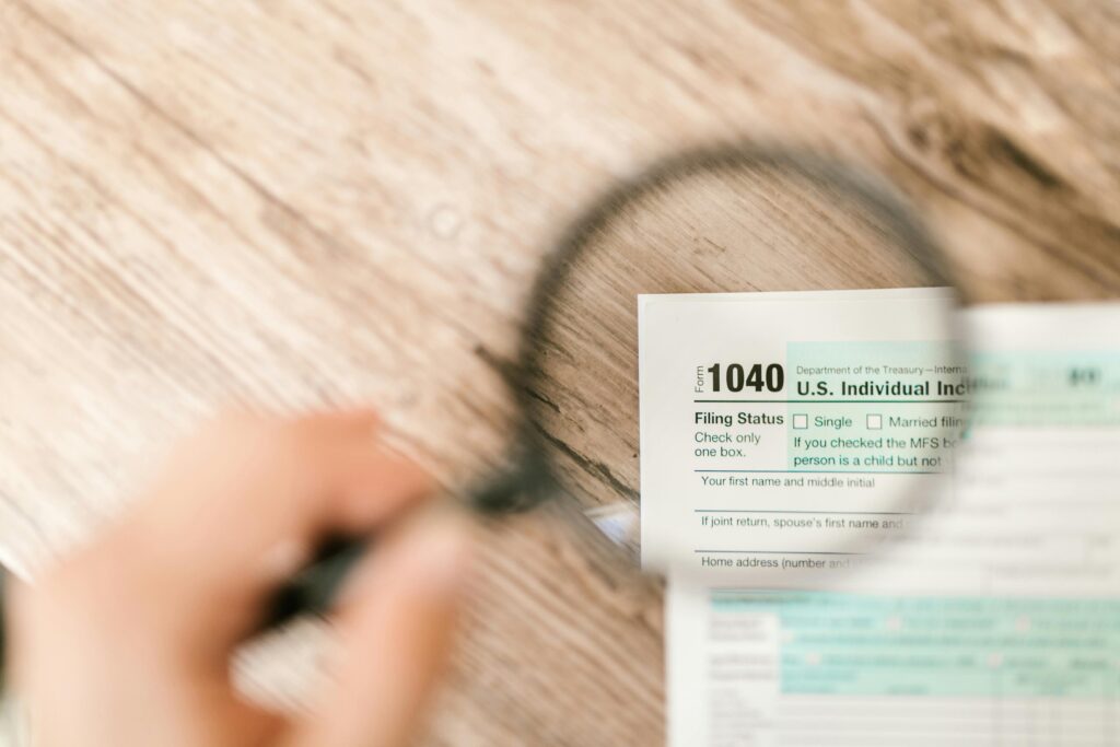 The upper left corner of an IRS 1040 tax form is shown through a magnifying lens. The form rests on a wooden surface.