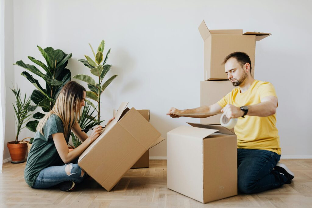 A white woman and man sit or kneel on a wooden floor with several moving boxes. The woman with long blonde hair is shown at left and wears a green T-shirt and ripped blue jeans. She uses a black market to write on a box. The white man to her right has short brown hair and a dark beard. He wears a yellow T-shirt, blue jeans and tennis shoes. He holds a roll of packing tape over the top folds of the box. In the background are 3 tall plants and multiple stacked boxes.