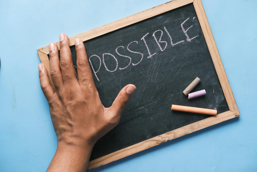 An African-American left hand hovers over a small chalkboard covering some letters (possibly IM), with the word POSSIBLE showing. Three pieces of chalk rest on the challkboard with a wooden frame. Background is light blue.