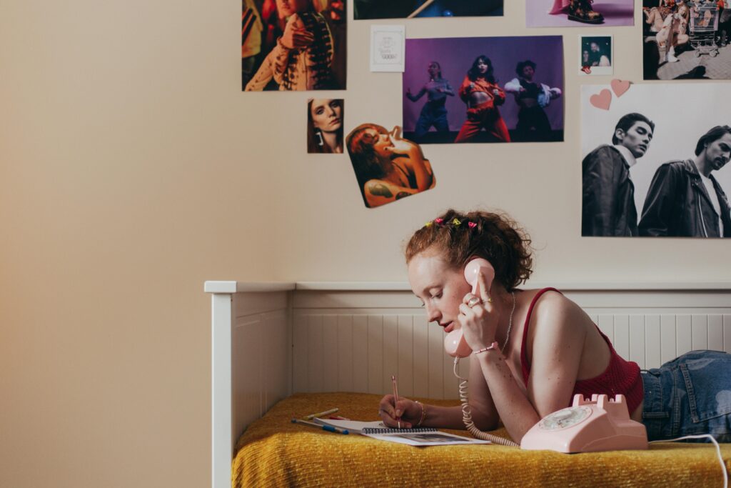 A young woman with red hair gathered on her head lays on a bed with a white headboard and golden yellow blanket. She wears a red camisole shirt and blue jeans. She is writing with a pink pen in a spiral bound book and holds the handset of a pink rotary phone in her left hand. On the wall in the background are posters and pictures of musical acts.