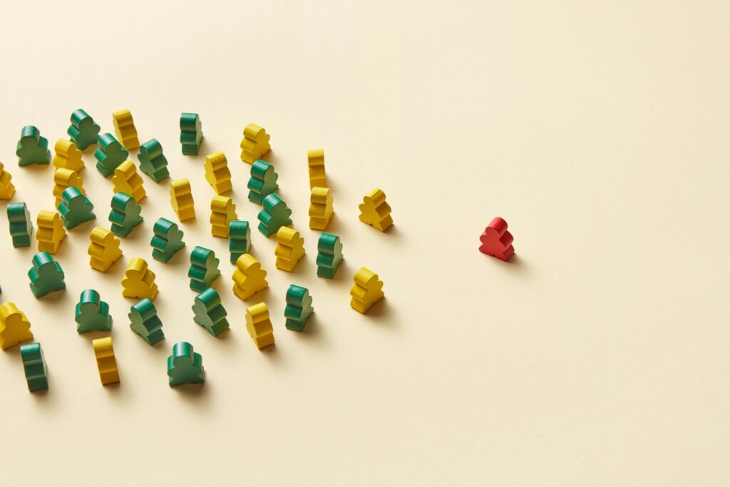 A grouping of yellow and green wooden figures shaped like a person is shown at the left. Standing solo a short distance from the grouping is a red wooden figure shaped like a person.