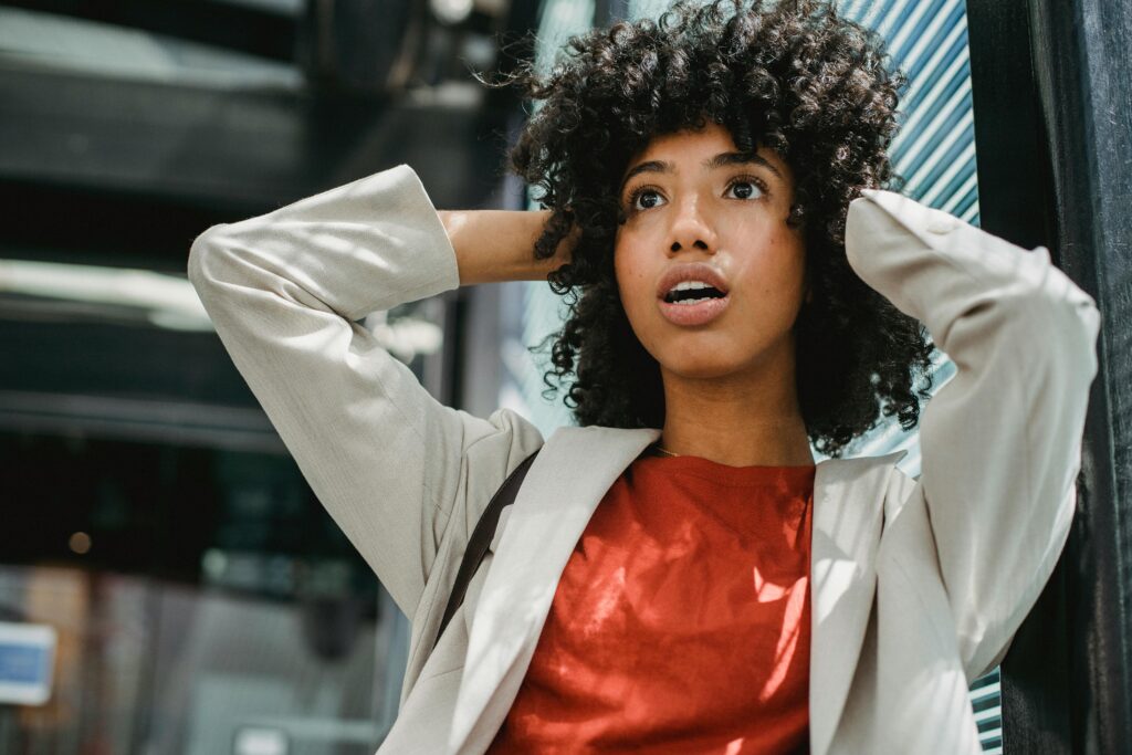 An African-American young woman with shoulder-length curly black hair is shown with a surprised look on her face and her arms raised and hands in her hair. She is wearing a burnt orange shirt and beige business jacket. The background seems to be a streetscape.