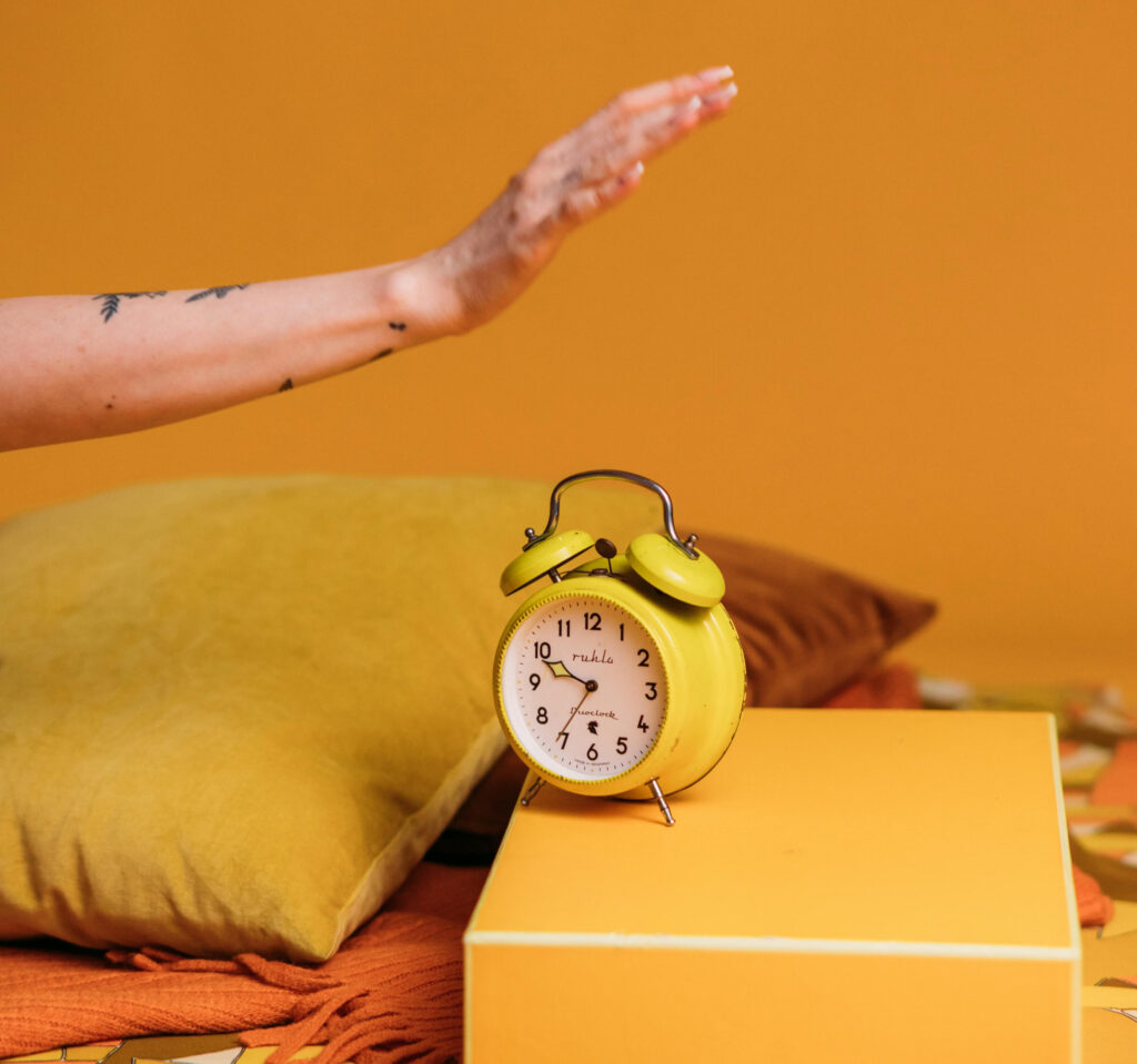 A woman's arm stretches into frame from the left and hovers over a bright yellow manual alarm clock sitting on a golden box stand. At left under the arm is a canary yellow pillow sitting on an orange blanket and a brown pillow is shown in the background.
