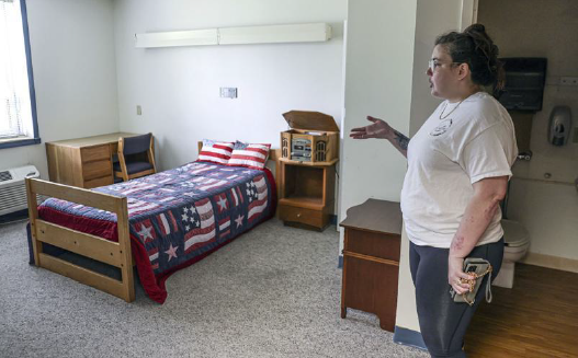 A young woman with dark hair, wearing a white T-shirt and black pants, gestures toward a room with a bed, desk and side table visible. On the bed are quilt and pillows with a U.S. flag motif. In the background, a bathroom can be seen.