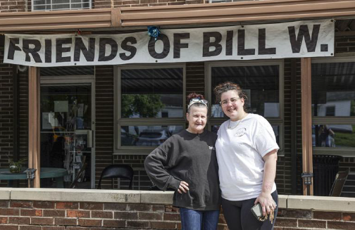 Two women stand in front of a brick building's patio area. The woman on the left has dark hair pulled back, and wears a dark grey sweatshirt and jeans. The woman on the right has dark hair, wears glasses, a white T-shirt and black pants. A sign saying "Friends of Bill W." hangs from the top of the patio.
