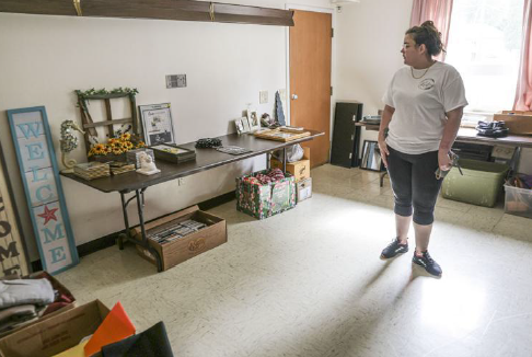 A woman stands in a room featuring a long dark table. She has dark hair pulled back and wears a white T-shirt, black capri pants and black shoes. Items that could be used to decorate an apartment - welcome signs, books and knick-knacks are displayed in the room.