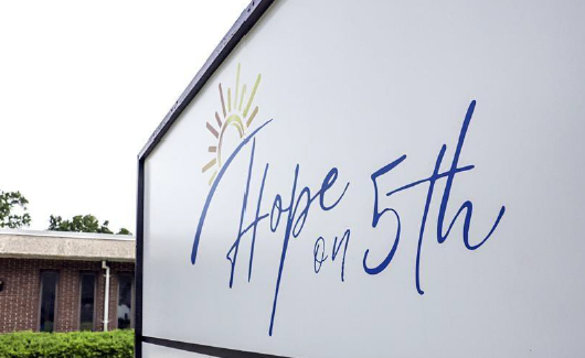 A white sign with the words "Hope on 5th" and featuring a rising sun is shown in the foreground. In the background is a brick building.