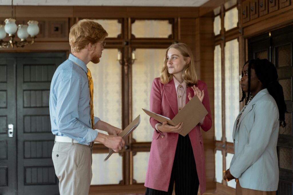 Three young professionals speak while standing in an entryway of an office. At left is a man with reddish hair, wearing glasses and a beard. He wears a light blue oxford shirt and light tan pants and holds a file folder. In the center is a woman with shoulder-length blonde hair. She wears a rose-colored business jacket, pink collared shirt and black pants, and also holds a file folder. At right is an African-American woman with long braids, wearing a white business jacket and tan pants.