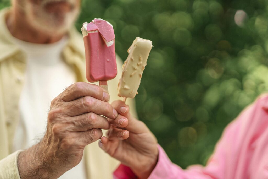 An older couple hold up ice cream desserts on sticks. On left, the man with a grey beard is shown in the background wearing a white T-shirt with yellow button-down shirt over it. The woman's hand is shown along with her pink shirt at right.