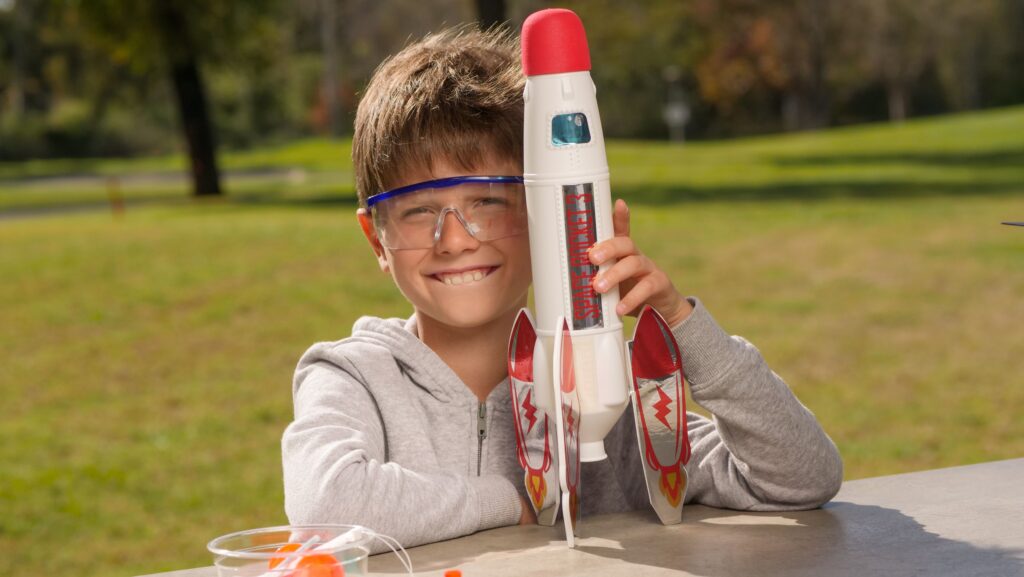 A smiling young boy with short light brown hair wearing safety glasses holds a model rocket. He appears to be in a park setting.