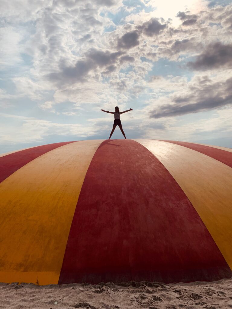A woman in silhouette wearing shorts and a tank top stands atop a yellow and red stiped dome along a beach. Clouds float in the blue sky in the background.