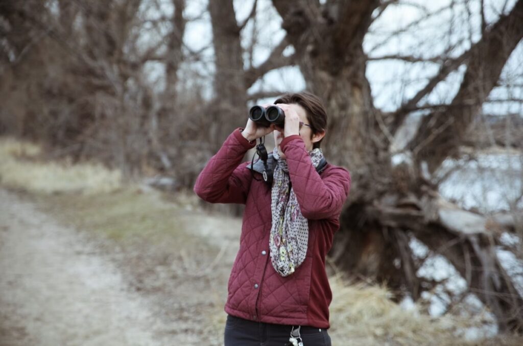 A woman with short dark hair holds a pair of binoculars to her face in a path that appears to run along a river (at right) in the fall with fallen trees in along the riverbank. She is wearing a maroon outdoors jacket with a gray scarf and black pants.