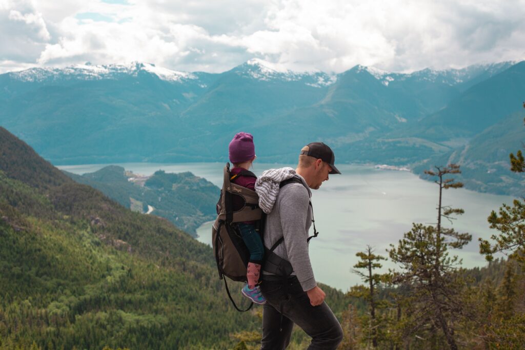 A father carries a toddler in a carrier on his back along a hiking trail surrounded by mountains and a lake in the background. The man has light hair and wears a black baseball cap, grey sweatshirt and black pants. The child wears a purple stocking cap, a fuschia sweater, blue pants, pink socks and blue sneakers.