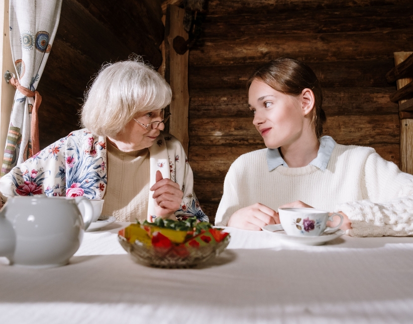 An elderly woman sitting looking at her granddaughter having a cup of tea.